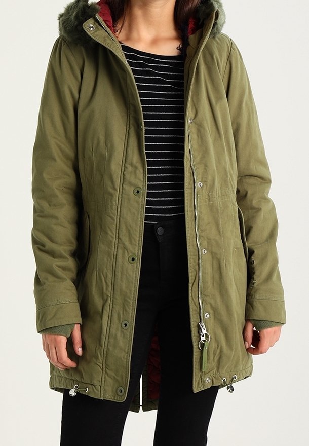 Femme portant un parka à capuche vert olive sur un chemisier rayé noir et blanc et un pantalon noir, debout devant un fond uni.