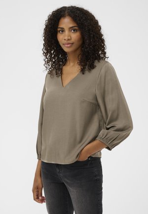 Blouse kaki à manches longues en tissu lisse avec un décolleté en V, des poignets froncés et une petite poche poitrine, assortie à un jean foncé.
