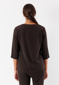 Tunica marrone con maniche a tre quarti, caratterizzata da un colletto rotondo e un orlo asimmetrico, realizzata in tessuto leggero e liscio.