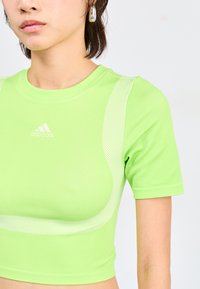 Žalias trumpų rankovių sportinis marškinėlis, pagamintas iš elastingo audinio. Turi išlenktą dizainą su dryžuotais akcentais ir iškilniuoju prekės ženklu ant krūtinės.