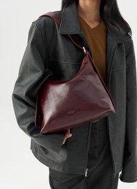 Persona che indossa una giacca di pelle nera e un top marrone, mentre tiene una borsa a tracolla in pelle rossa scura con fibbia argentata.