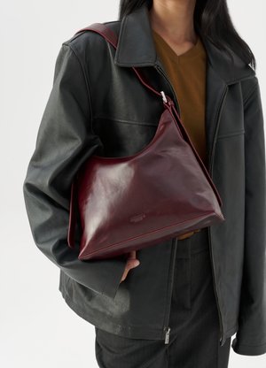 Personne portant une veste en cuir noir et un haut marron, tenant un sac à épaule en cuir rouge foncé avec une boucle en argent.