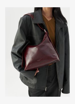 Personne portant une veste en cuir noir et un haut marron, tenant un sac à épaule en cuir rouge foncé avec une boucle en argent.