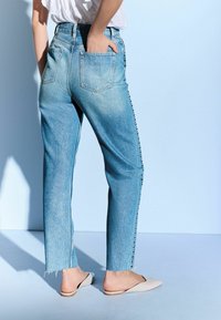 Jeans de culoare albastru deschis, cu talie înaltă și picior dreapt, textură estompată și butoane pe lateral. Asortați cu pantofi slip-on bej, pe un fundal minimalist.
