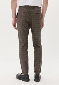 Braun karierte Hose mit taillierter Passform, ausgestattet mit Taschen mit Knopfverschlüssen, cropped Länge und glatter Textur. Kombiniert mit dunklen Schuhen.