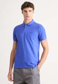 Camisa polo azul de mangas curtas, feita de algodão, com uma pala de três botões e um logótipo discreto no peito. Usada com calças cinzas.
