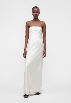 Femme portant une robe de satin blanche sans bretelles, longue jusqu'au sol, et des talons ouverts noirs, debout devant un fond clair uni.