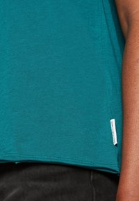 Camicetta teal a maniche corte in morbido tessuto, con etichetta laterale e vestibilità rilassata con orlo dritto. Sfondo nero visibile sotto.