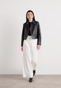 Een vrouw loopt zelfverzekerd in een zwart leren jack, zwarte top en wijde witte broek, tegen een minimalistische achtergrond.