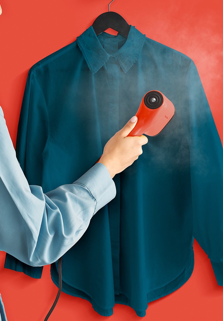 Stoomkledingreiniger in het rood, gebruikt op een teal hangend shirt. Heeft een gladde textuur, ronde spuitmond en stoom die wordt uitgestoten tegen een rode achtergrond.