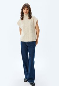 Crème kleurige kabelgebreide mouwloze trui met een hoge kraag, gecombineerd met wijduitlopende donkerblauwe denim jeans en zwarte schoenen met rafelige accenten.