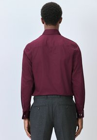 Bordeauxrotes Langarmhemd mit Kentkragen und taillierter Passform, kombiniert mit grauen maßgeschneiderten Hosen mit Seiten- und Gesäßtaschen.