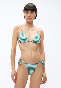 Žena v modrozeleno-bielych pruhovaných textúrovaných bikinách so zaväzovaním na bokoch, stojaca pred jednoduchým bielym pozadím.