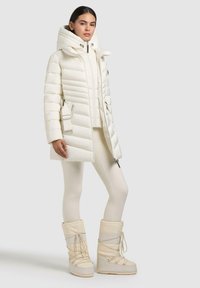 Crèmekleurige pufferjas met capuchon, ritssluiting aan de voorkant en zijzakken. Gecombineerd met crème legging en hoge sneeuwlaarzen met veters.