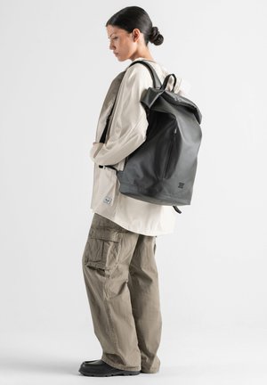 Persona joven con el cabello oscuro recogido lleva una chaqueta beige, pantalones cargo holgados color caqui, zapatos negros y una mochila grande negra, de pie de perfil.
