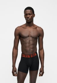 Μυώδης άνδρας που φοράει μαύρα μποξεράκια Calvin Klein με πορτοκαλί ελαστική ζώνη με κείμενο, μπροστά σε απλό γκρι φόντο.