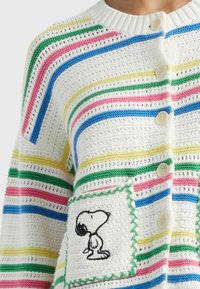 Cardigan tricoté en blanc, avec des rayures horizontales vertes, bleues, roses et jaunes. Inclut un motif Snoopy brodé sur une poche.