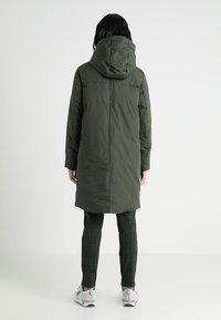 Abrigo largo acolchado en verde oliva con capucha, de corte recto, con costuras visibles y bolsillos laterales. Llevado sobre pantalones de cuadros y zapatillas deportivas.