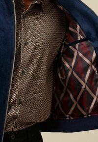 Granatowa zamszowa kurtka bomberka rozpięta, odsłaniająca brązową koszulę w wzory oraz kontrastowy geometryczny wzór podszewki w bordo i czarnym kolorze.