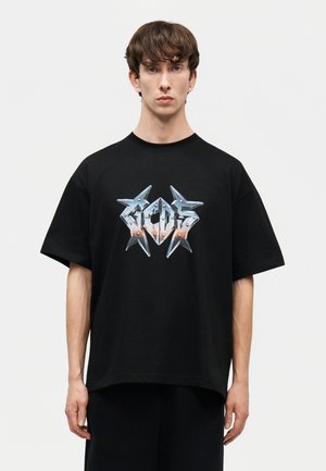 Νεαρός άνδρας με κοντά καστανά μαλλιά, φορώντας μια φαρδιά μαύρη μπλούζα με ασημένιο λογότυπο "GCDS" με αγκάθια, πάνω σε απλό λευκό φόντο.