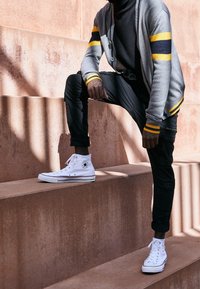 Cardigan gris avec des rayures jaunes et bleu marine, pantalon noir et baskets montantes blanches, posée sur des marches à surface texturée.