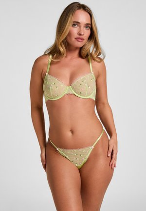 Frau trägt hellgrünen transparenten BH und passenden Tanga mit Blumenstickerei vor einfarbig grauem Hintergrund.