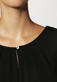 Gros plan sur le cou et le haut de la poitrine d'une personne portant un haut noir fermé par un petit bouton blanc unique.