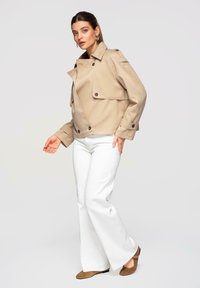 Femme portant une veste trench courte beige, un pantalon blanc ample et des chaussures en daim marron, posant devant un fond clair uni.