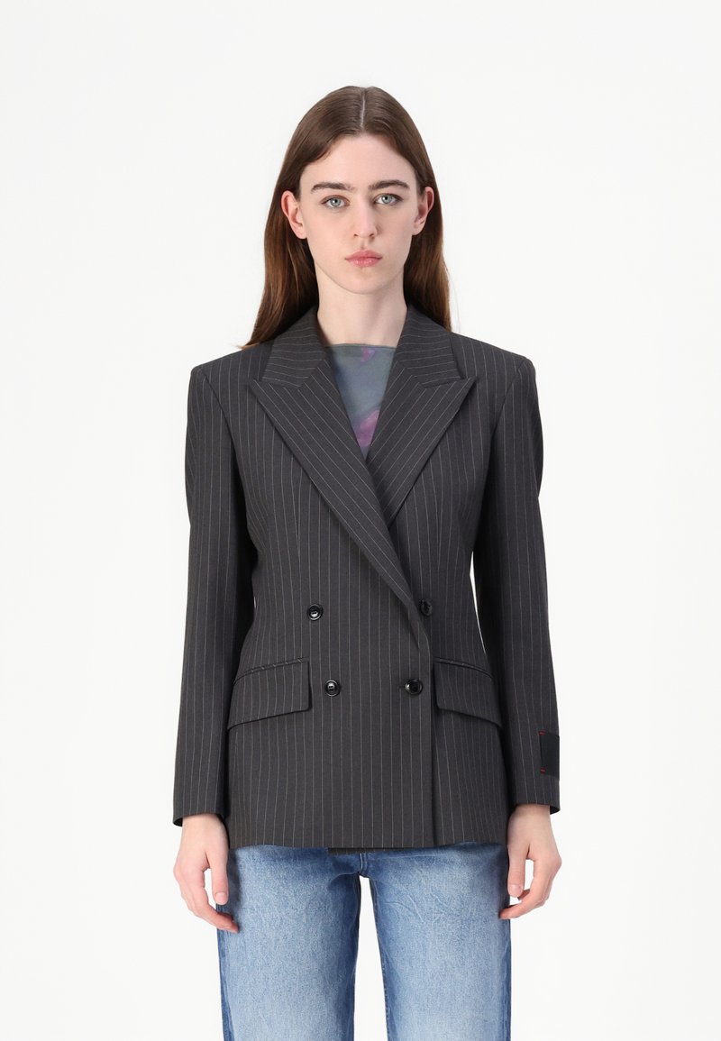 Blazer gris à rayures fines croisé avec larges revers, deux poches avant et boutons noirs. Porté sur un haut coloré et associé à un jean bleu.