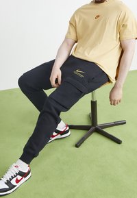 Nike beige T-shirt med logotyp, kombinerad med marinblå cargo-byxor med fickor och gul branding. Svarta, röda och vita sneakers fulländar outfiten.