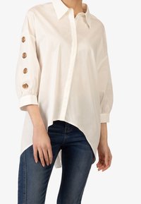 Camicia bianca con colletto a punta, maniche lunghe con bottoni dorati e orlo asimmetrico, abbinata a jeans blu.