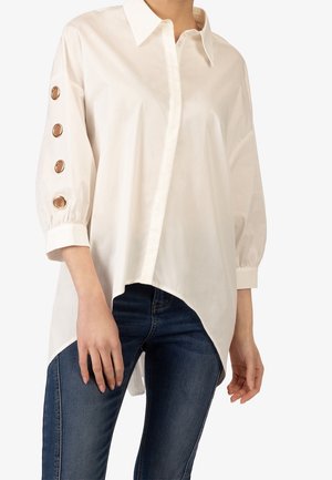 Chemise blanche à col pointu, à manches longues avec des boutons dorés, et un ourlet asymétrique, associée à un jean bleu.
