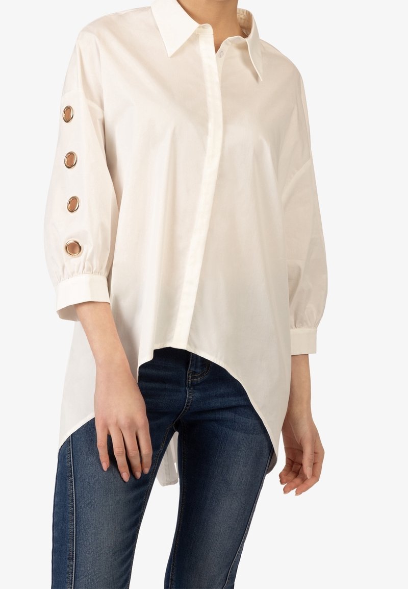 Camicia bianca con colletto a punta, maniche lunghe con bottoni dorati e orlo asimmetrico, abbinata a jeans blu.