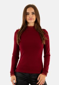 Camicetta a maniche lunghe color bordeaux a costine con colletto alto e polsini arricciati, caratterizzata da bottoni decorativi sui polsi. Tessuto liscio, design aderente.