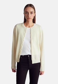 Kremowy kardigan z miękkiej dzianiny, z okrągłym dekoltem i zapięciem na guziki, noszony na białej koszuli i połączony z czarnymi dżinsami.