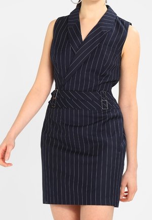 Femme portant une robe bleu marine sans manches à rayures fines avec une large ceinture et un col à revers.