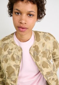 Giubbotto bomber trapuntato tan con motivo camouflage sopra una camicia rosa chiaro, caratterizzato da bottoni a pressione e un collo rotondo. Texture liscia.