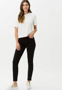 Witte t-shirt met korte mouwen gecombineerd met zwarte skinny jeans. Model draagt lichtgekleurde sneakers. Glad materiaal en aansluitend ontwerp.