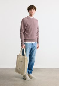 Νεαρός με κατσαρά μαλλιά που φοράει πουλόβερ πλεκτό σε μωβ απόχρωση, ανοιχτόχρωμα μπλε τζιν, μπεζ αθλητικά παπούτσια και κρατάει μια μπεζ τσάντα χειρός με μπροστινή τσέπη.