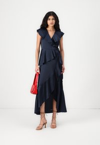 Vestido de cetim navy com detalhes em babados, bainha assimétrica e design envolvente. Combinado com uma bolsa vermelha e sandálias bege com tiras.