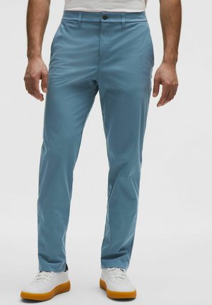 Mann trägt hellblaue, schmal geschnittene Hose und weiße Sneaker mit braunen Sohlen, steht vor einem schlichten weißen Hintergrund.