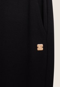 Pantaloni jogger neri in tessuto, caratterizzati da una tasca laterale e un emblema quadrato in rosé oro lucido con la scritta "FY". Texture liscia con dettagli minimi.