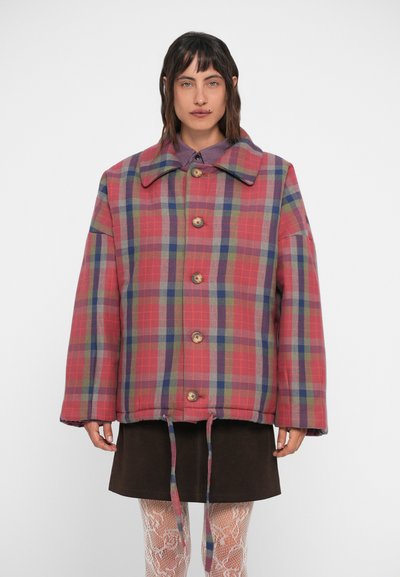 Veste rouge à carreaux avec un grand col, des boutons en bois et un lien à l'avant. Portée sur une jupe marron et des collants à motifs.