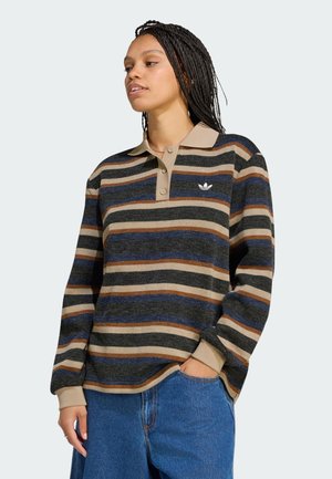 Woman wearing a long-sleeve striped polo shirt in beige, navy, and brown with blue jeans, standing with one hand in her pocket.