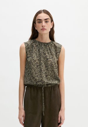 Femme aux longs cheveux bruns portant un haut sans manches vert à fleurs et un pantalon ample marron foncé sur un fond clair uni.