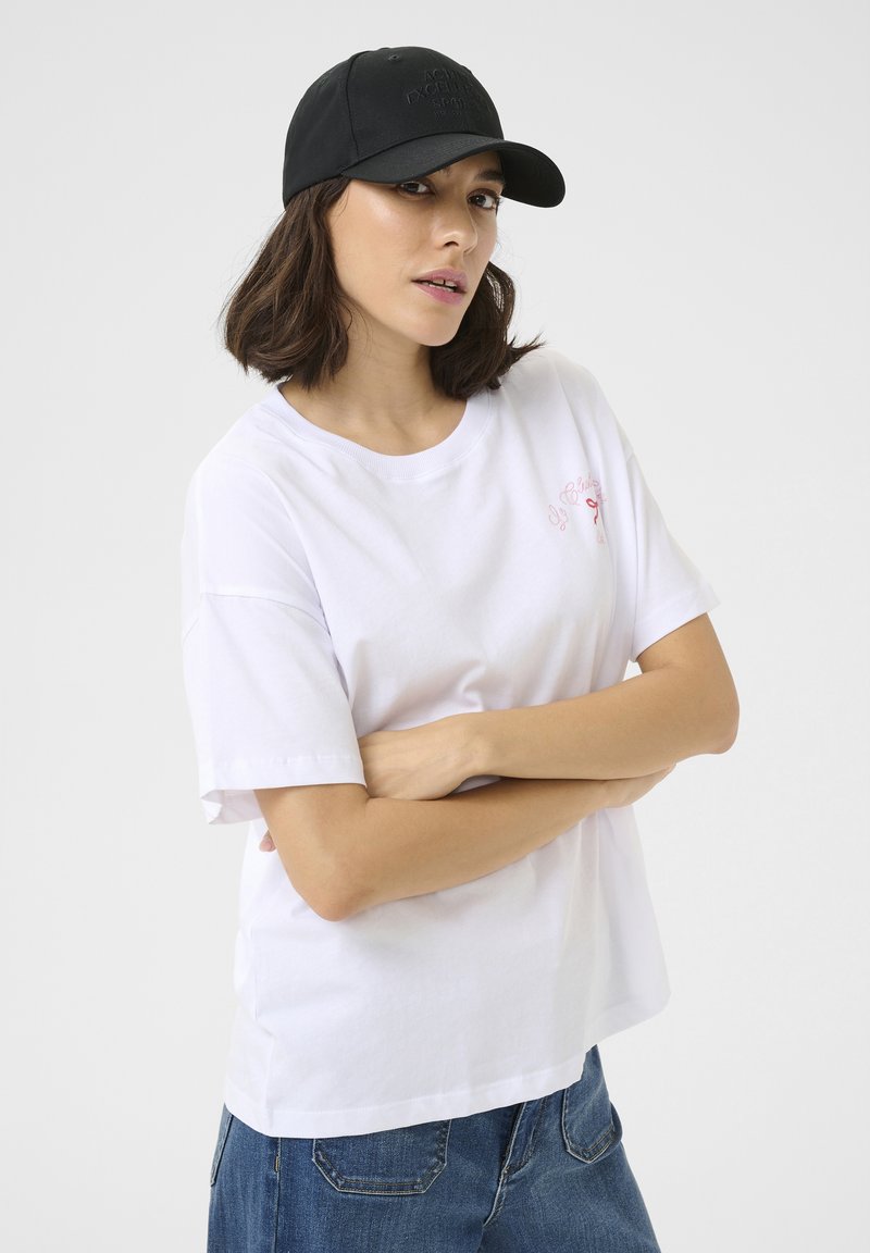 Camiseta de algodón blanca con mangas cortas, con un pequeño gráfico rosa en el pecho. Combinada con una gorra negra ajustable.