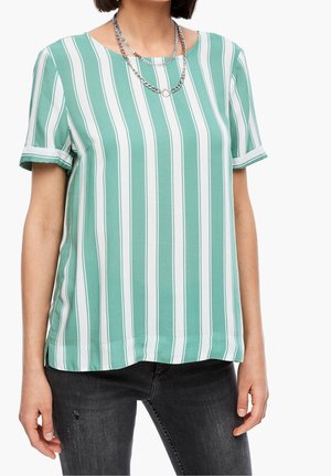 Femme portant un haut à manches courtes à rayures verticales vertes et blanches, un jean noir usé et des colliers en chaînes argentées superposées.