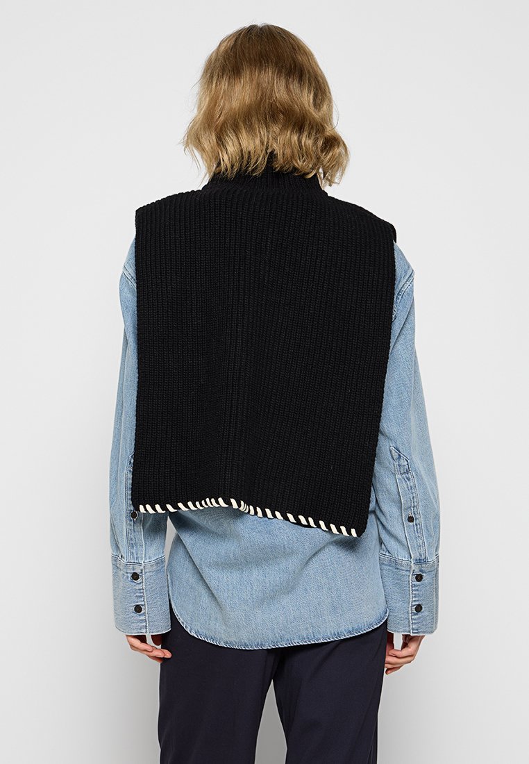 Schwarze gestrickte Weste mit gerippter Textur, featuring einen gestreiften Rand, getragen über einem hellblauen Jeanshemd. Rückansicht, die die Länge und Form des Kleidungsstücks zeigt.