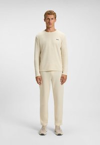Haut beige à manches longues et pantalons assortis, tissu texturé, design ajusté, petit logo noir sur la poitrine, associé à des baskets beiges.