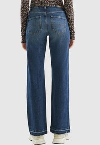 Weite blaue Jeans mit einem verwaschenen Textur, Fünf-Taschen-Design und ausgefransten Säumen. Getragen mit weißen Sneakern und einem blumigen langärmeligen Oberteil.